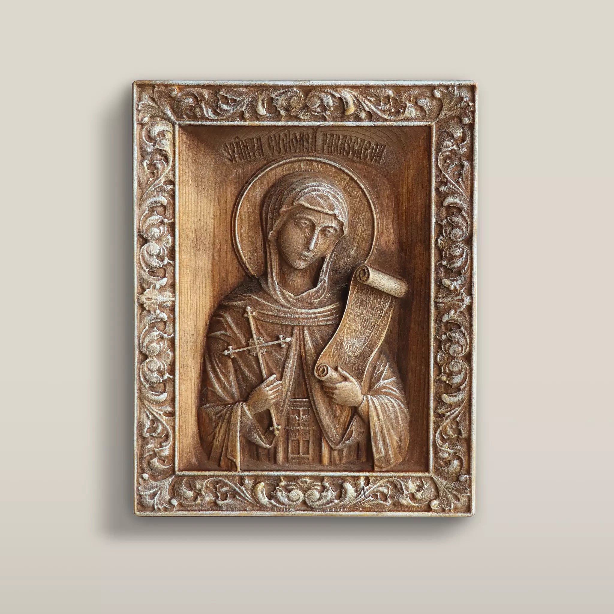 Carved wooden icon of Saint Paraskeva holding a cross and an open scroll, set within a carved leaf border