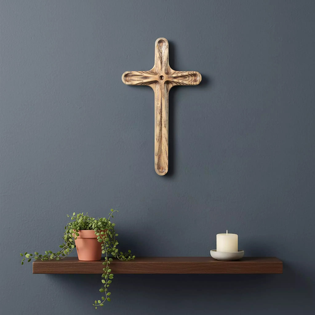 The cross hung on a dark blue wall above a floating shelf with a plant and a candle