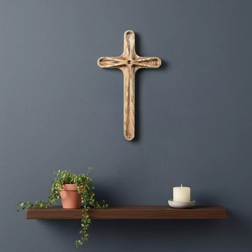 The cross hung on a dark blue wall above a floating shelf with a plant and a candle